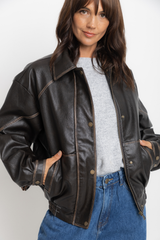 Eddie vegan leather jacket in chocolate, oversized bomber-style jacket with snap buttons, styled with a grey sweater, plaid shirt tied at the waist, and denim jeans