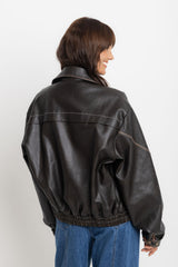 Eddie vegan leather jacket in chocolate, oversized bomber-style jacket with snap buttons, styled with a grey sweater, plaid shirt tied at the waist, and denim jeans