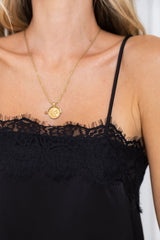 A close-up of a model wearing a delicate gold chain necklace with a round, textured gold pendant, styled with a black lace-trimmed camisole top.