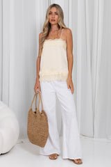A woman stands in a studio wearing a cream spaghetti-strap camisole top with lace trim, white wide-leg pants, and holding a woven straw tote bag.