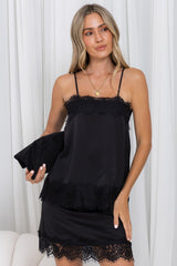 A woman wearing a black satin slip dress with lace trim and holding a matching black clutch bag, posed in front of white curtains.