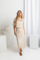A model wearing a crochet top and midi-length beige crochet skirt with a ribbed elastic waistband, in a fitted silhouette, paired with a crochet top and brown sandals.