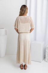 A model wearing a crochet top and midi-length beige crochet skirt with a ribbed elastic waistband, in a fitted silhouette, paired with a crochet top and brown sandals.