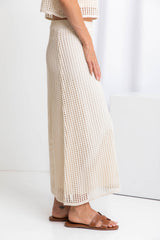 A model wearing a midi-length beige crochet skirt with a ribbed elastic waistband, in a fitted silhouette, paired with a crochet top and brown sandals.