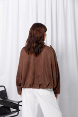 Woman wearing a brown suede-look jacket over a white top and white pants, standing against a white curtain background.