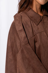 Woman wearing a brown suede-look jacket over a white top and white pants, standing against a white curtain background.