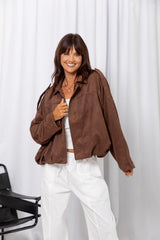 Woman wearing a brown suede-look jacket over a white top and white pants, standing against a white curtain background.