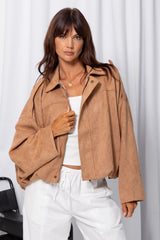 Woman wearing a sand suede-look jacket over a white top and white pants, standing against a white curtain background.
