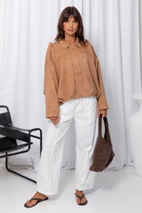Woman wearing a sand suede-look jacket over a white top and white pants, standing against a white curtain background.
