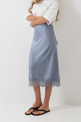A model wearing a light blue midi skirt with a lace hem, paired with a white top and black flip-flops, standing against a plain white studio background.