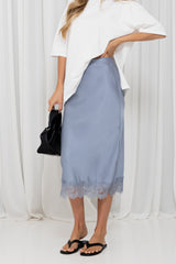 A model wearing a light blue midi skirt with a lace hem, styled with an oversized white t-shirt, black flip-flops, and holding a black handbag against a white curtain backdrop.