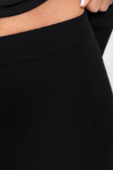 Close-up of a black ribbed knit garment, likely a skirt or pants, worn by a model. The image shows the waistband, ribbed texture, a portion of the model's skin at the top, and a hand with white nail polish at the upper right edge.