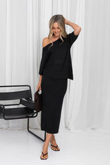 A woman models a black two-piece outfit consisting of an asymmetrical off-the-shoulder top and a matching midi skirt, standing in front of white draped curtains.