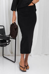 A model wearing a black midi-length bodycon skirt, styled with a black top, brown sandals, and holding a brown woven clutch bag, set against a white curtain background.