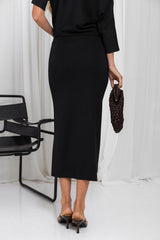 A woman wearing a black midi skirt and black top, holding a dark brown woven clutch, standing in front of white curtains.