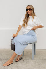 A woman wearing a white oversized t-shirt and a light blue midi skirt sits on a gray stool, holding a small black woven handbag. She is wearing dark sunglasses and black flip-flop sandals against a plain white wall and concrete floor.
