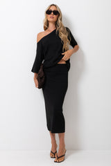 A woman modeling a black one-shoulder midi dress with a side cutout, styled with black sunglasses, a brown woven clutch, and black strappy heels against a plain white wall.