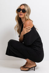 A woman crouching in a black off-the-shoulder jumpsuit with wide-leg pants, wearing black sunglasses and brown heeled thong sandals against a plain white studio background.