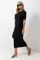 A woman modeling a black short-sleeve top and matching midi skirt set against a plain white studio background. She is wearing black sunglasses, black strappy heels, and holding a small dark woven handbag.