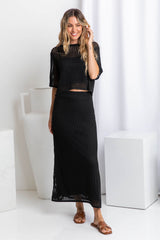 A model wearing a crochet knit black top with a midi length black crochet knit skirt with a ribbed elastic waistband and fitted silhouette.