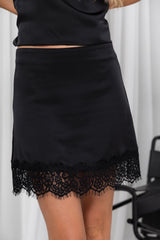 A black satin mini skirt with a lace hem, shown on a model wearing a matching black top. The background is a white curtain.