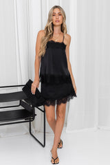 A woman models a black satin slip dress with lace trim, holding a matching black clutch bag in a bright studio setting with white drapes.