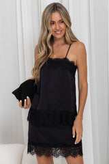 A woman wearing a black spaghetti-strap slip dress with lace trim, holding a matching black clutch bag against a white curtain background.