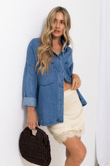 A woman models a blue denim button-up shirt with rolled sleeves, worn open over a cream satin crop top and white lace-trimmed shorts, holding a dark brown woven handbag against a white background.