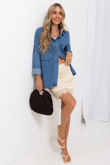 A woman models a blue denim button-up shirt with rolled sleeves, paired with a cream lace-trimmed mini skirt and white strappy heels, holding a dark brown woven handbag against a white studio backdrop.