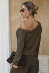 Woman wearing a brown off-shoulder sweater and holding a clutch in an indoor setting.