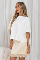 A woman wearing a white short-sleeve crop top and a cream satin mini skirt with a lace hem, standing in front of white curtains.