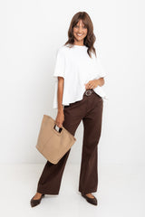 Woman wearing a white top and brown pants holding a beige handbag on a white background