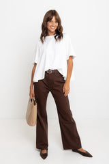 Woman wearing a white blouse and brown pants with a beige bag against a white background