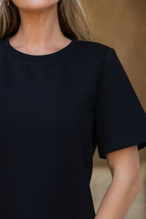 Close-up of a person wearing a plain black short-sleeve crew-neck top against a softly blurred warm brown background.