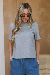 A woman wearing a light heather gray short-sleeve crewneck t-shirt and blue drawstring pants, standing against a warm beige textured wall.