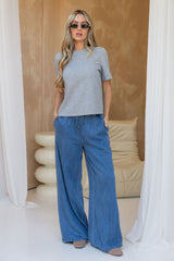 A woman modeling a short-sleeve heather gray t-shirt and wide-leg blue denim pants, standing in a minimalist indoor setting with draped white curtains and a beige wall.
