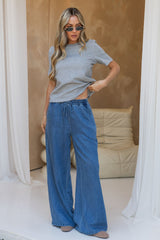 A woman modeling a light gray short-sleeve knit top and wide-leg blue denim pants in a bright indoor setting with draped white curtains and a beige lounge chair in the background.