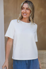 A woman wearing a white short-sleeve crewneck top and blue jeans, standing against a warm beige textured wall.