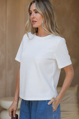 A woman wearing a white short-sleeve crewneck t-shirt and blue jeans, standing against a neutral beige background.