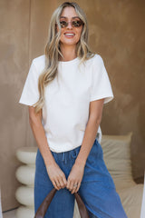 A woman wearing a white short-sleeve crewneck t-shirt and blue jeans, standing in front of a textured beige wall.