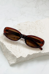 Oval sunglasses with a translucent dark brown acetate frame and dark brown lenses, resting on a textured white stone block over a light marble surface.