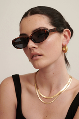 A close-up of a model wearing tortoiseshell cat-eye sunglasses, a layered gold necklace, and a gold hoop earring against a plain light beige background.
