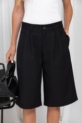 A person wearing black wide-leg cropped trousers with a button waistband and front pleats, paired with a white top. The model is holding a black handbag against a light neutral indoor background.