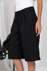 A model wearing black wide-leg pleated shorts with a button closure, styled with a white top and holding a black handbag against a white studio backdrop.