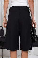 A person wearing black wide-leg cropped shorts or culottes with a white top, holding a black handbag in a bright indoor setting.