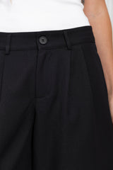 Close-up of a person wearing black pleated trousers with a button waistband, paired with a white ribbed top.