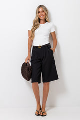 A woman modeling a white short-sleeve top and black knee-length shorts with a gold-tone belt, holding a dark brown woven handbag against a plain white studio background.