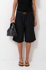 A model wearing black pleated knee-length shorts with a matching black belt featuring a gold buckle, styled with a black top, black sandals, and holding a black handbag against a plain white background.