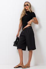 A woman modeling a black short-sleeve crop top with a side cutout, paired with black pleated knee-length shorts, a black belt with a gold buckle, black sandals, and holding a black handbag against a plain white studio background.
