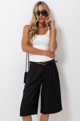 A woman modeling black pleated culotte shorts with a black belt, styled with a white tank top, black sunglasses, and a small black shoulder bag against a plain white background.